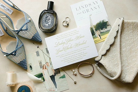 Wedding invitation suite flat lay with save the date and engagement photo card, rings, perfume, blue heels, pearls on neutral fabric backdrop