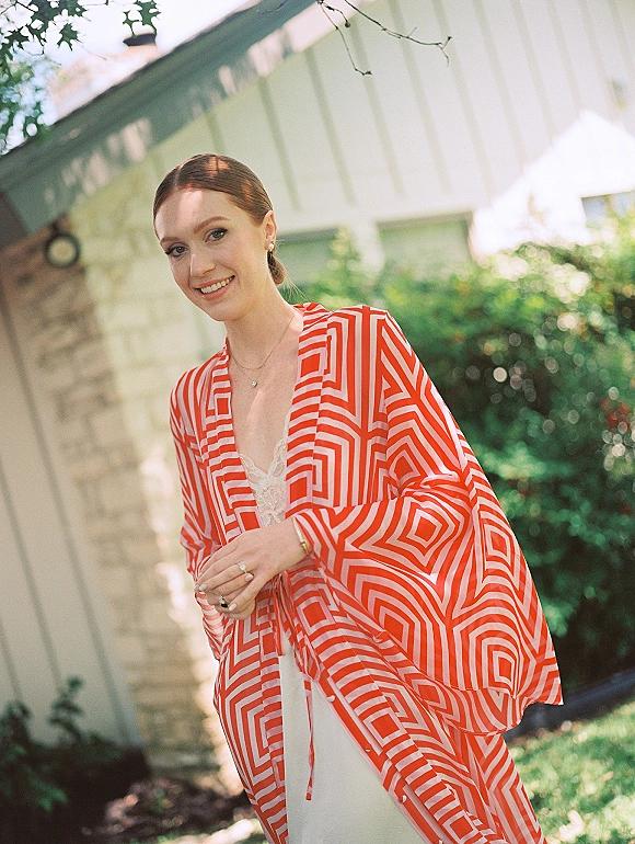 Bride getting ready in a patterned robe over a lace camisole, pearl earrings and rings, standing in sunlight by a house exterior