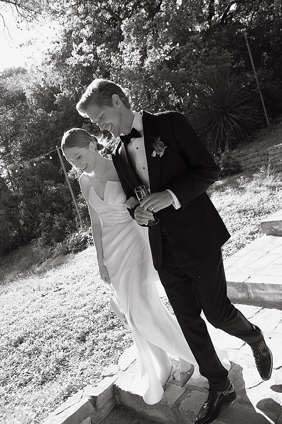 Wedding couple portrait of bride in a birdcage veil and strapless dress walking arm in arm with groom holding a champagne flute on sunlit garden stone steps