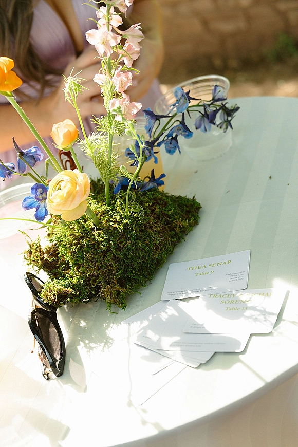 Escort cards display with wedding escort cards arranged beside a moss centerpiece and ranunculus blooms on a sunlit white table by a stone wall