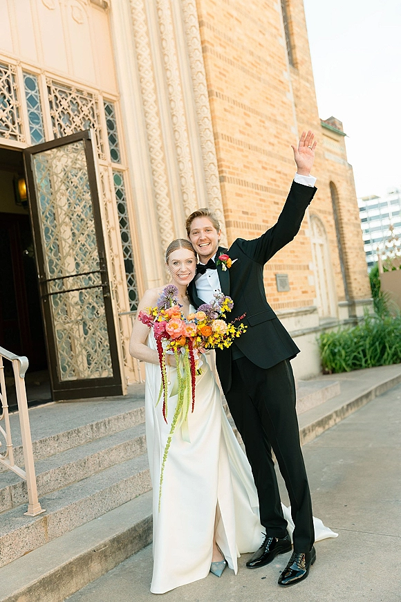 Couple portrait of bride and groom outside church, bride in satin gown with veil and bouquet, groom in black tux waving on steps
