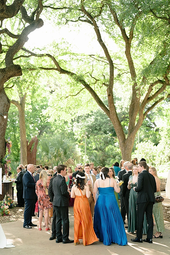 Wedding cocktail hour with guests mingling outdoors, holding cocktail glasses in suits and long dresses under a tree canopy near a stone wall