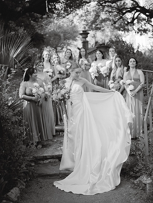Bride with bridesmaids walking down stone steps, bride holding bouquet and veil with floral accents, bridesmaids in long dresses amid garden foliage