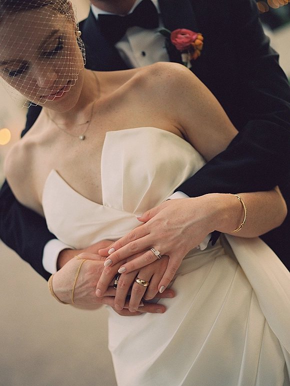Couple portrait of groom hugging bride, showcasing birdcage veil and wedding rings against soft bokeh lights on an outdoor walkway