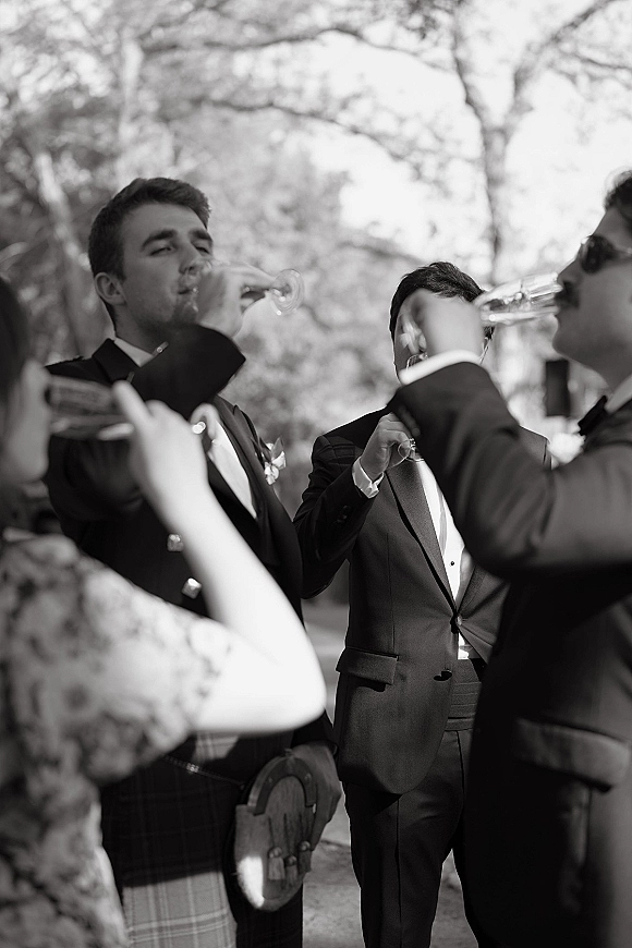 Groomsmen toast with champagne flutes, tuxedos and bow ties, some in sunglasses, sharing laughs outdoors among leafy trees