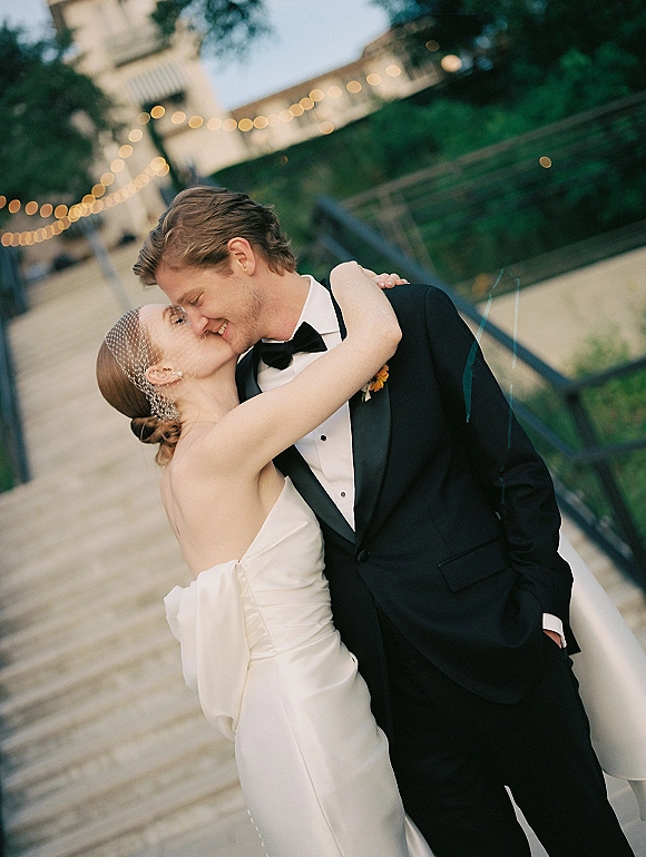 Wedding kiss portrait of bride and groom kissing under string lights on an outdoor walkway, her birdcage veil and strapless dress at twilight