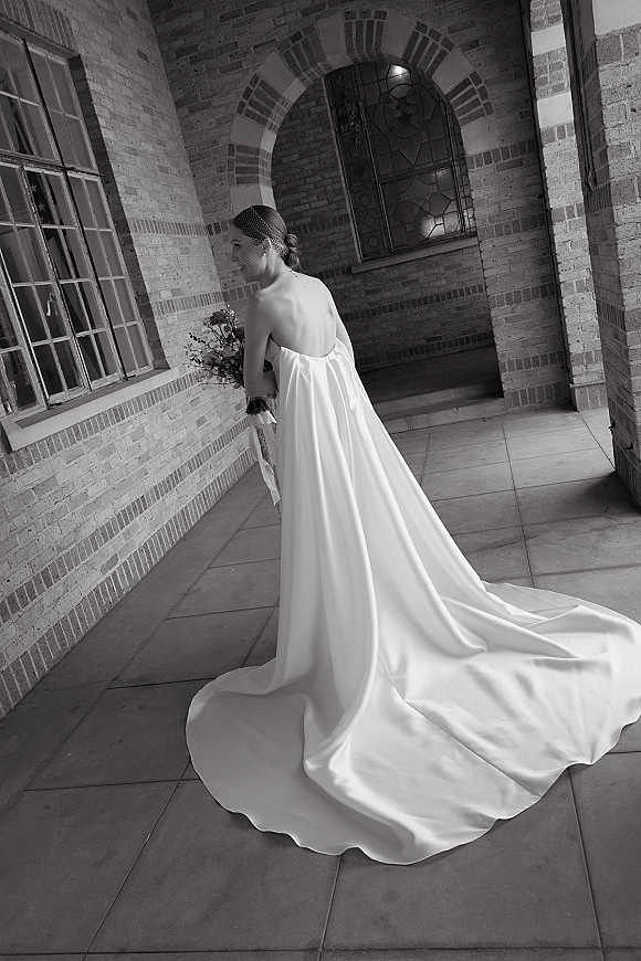 Bridal portrait of a bride in a strapless wedding dress with a long train, holding a bouquet under a brick archway corridor