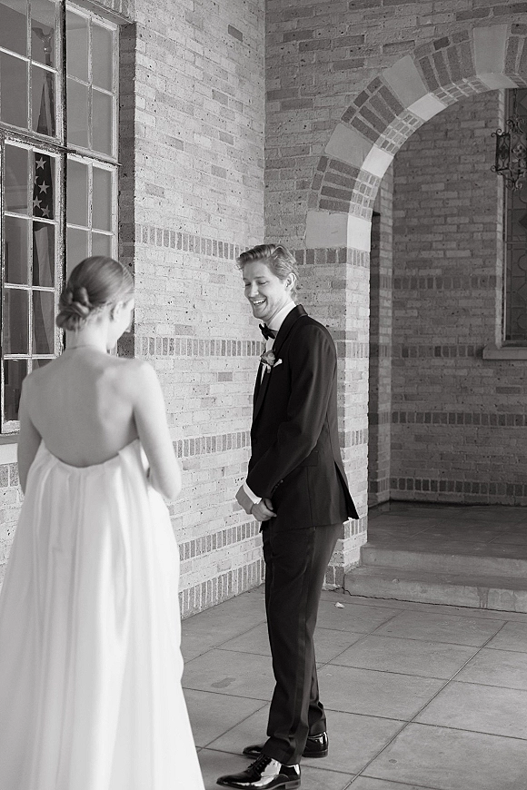 First look moment as bride in a wedding dress approaches groom in tuxedo with bow tie and boutonniere by a brick arched doorway
