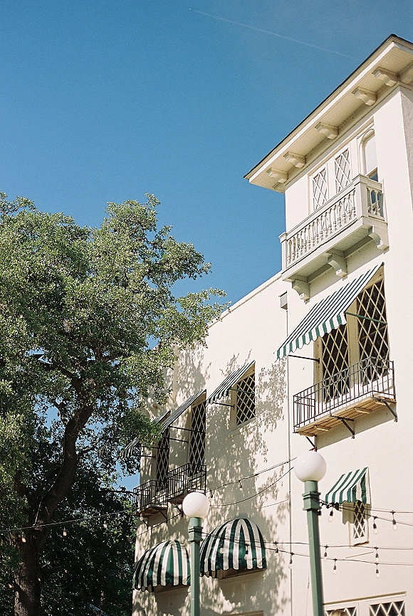 Wedding venue exterior with striped awnings and balcony, framed by leafy trees under a blue sky, accented with string lights and lanterns