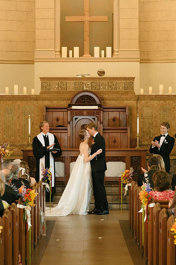 Ceremony kiss as bride in a strapless gown and groom in tuxedo kiss at the church altar with wood cross, candles, and guests in pews
