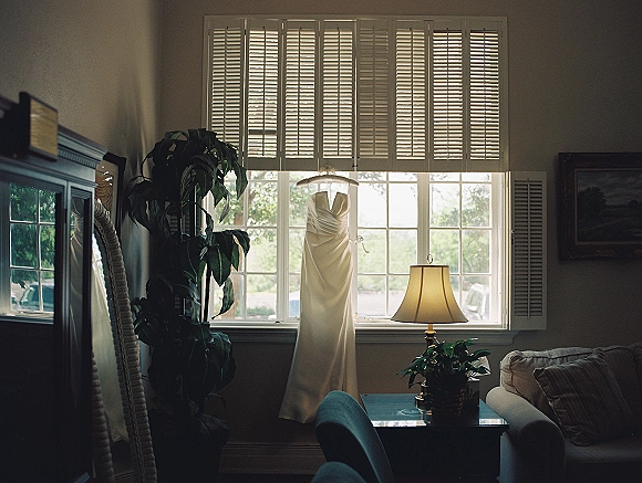 Wedding dress hanging in a bright window, strapless satin gown on a hanger with a soft table lamp nearby in a cozy room