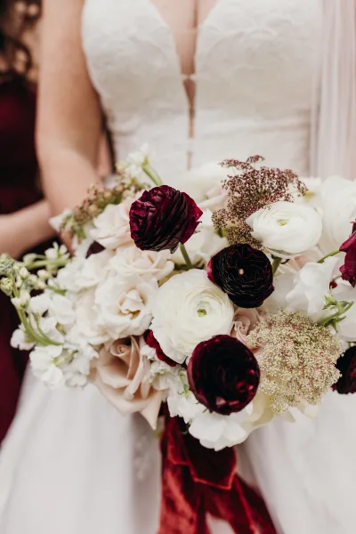 Bridal bouquet of burgundy and white bouquet with white roses and ranunculus, greenery, and trailing ribbon held against the bride’s bodice