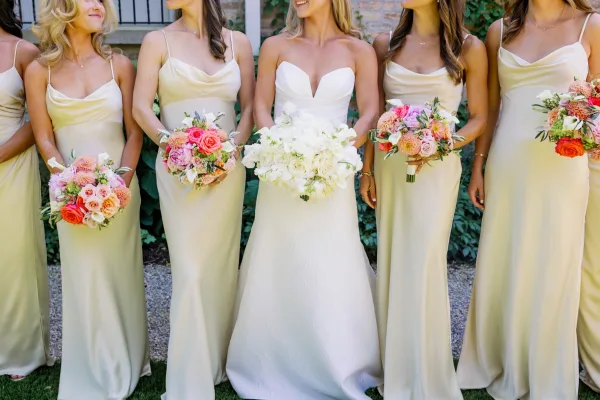 Bride and bridesmaids in champagne bridesmaid dresses smile, holding coral and pink bouquets against garden greenery and a brick wall.