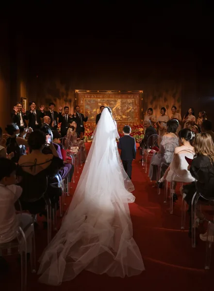 Wedding processional as bride walking down aisle in a cathedral veil and long train on a red runner, with guests seated and floral arrangements