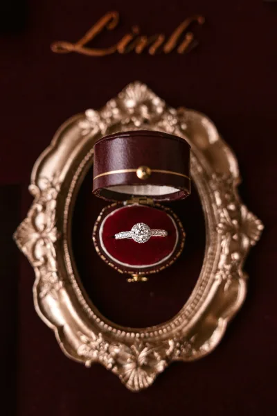 Engagement ring with a diamond halo resting in a burgundy velvet ring box with gold trim, styled beside an ornate gold frame backdrop
