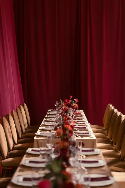 Reception tablescape with a long banquet table setup, red floral centerpiece, place cards and glassware against burgundy drapery backdrop indoors
