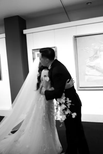 Wedding first look as bride and groom hug in a hallway, her lace dress and long veil trailing behind his tuxedo with boutonniere