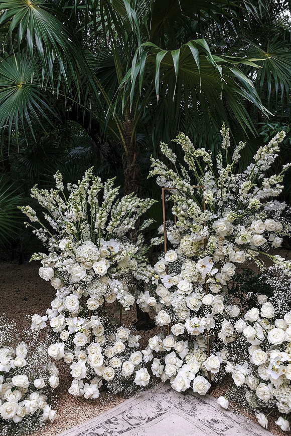 Wedding ceremony flowers in a white floral ceremony arrangement with roses, orchids, and hydrangeas on gold stands in a palm garden setting