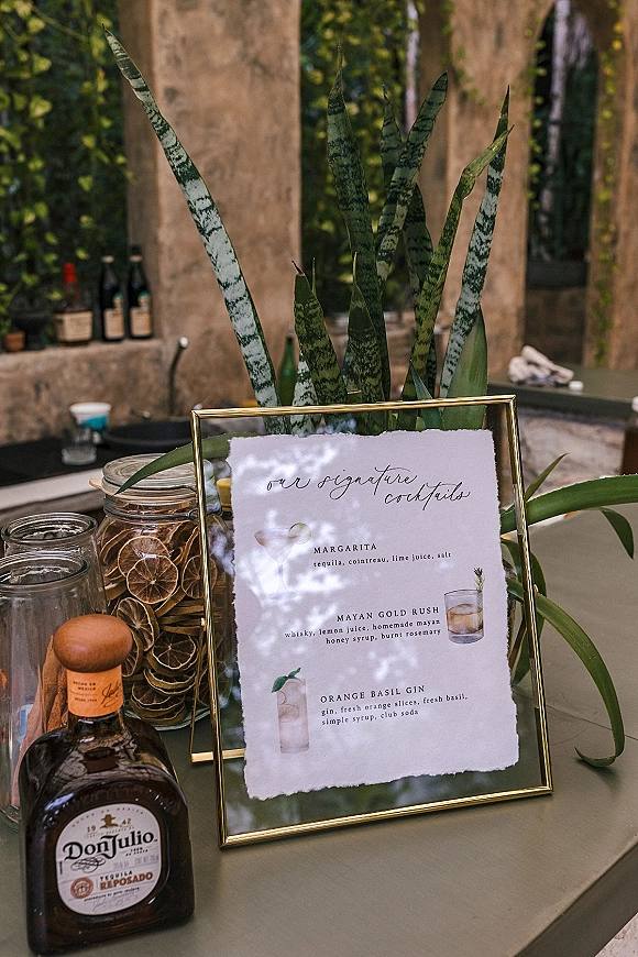 Signature cocktail sign in a gold frame on a wedding bar table with tequila, glassware, and dried citrus jars against a stone wall with greenery