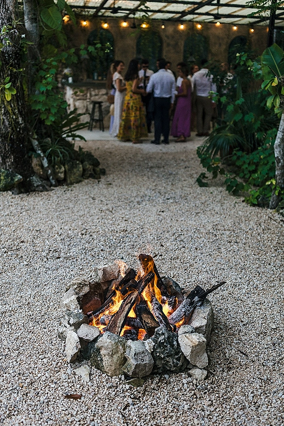 Wedding fire pit with glowing flames in a stone ring, outdoor wedding fire pit under string lights beside a covered bar in a gravel courtyard