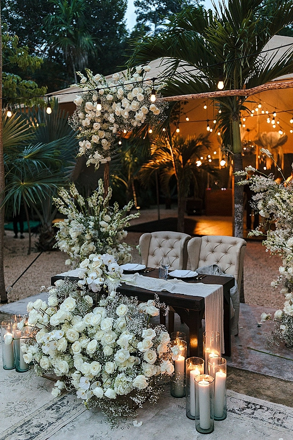 Reception sweetheart table with sweetheart table decor of white roses, orchids, candles in glass cylinders under string lights in a garden tent