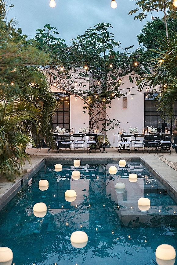 Poolside reception decor with string lights and floating pool lights illuminating long tables, candles, and white florals beside a pool at dusk