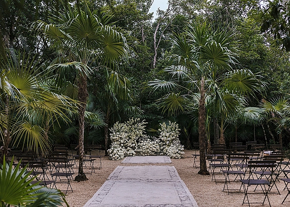 Ceremony setup with outdoor ceremony seating, layered aisle runner rugs leading to a white floral installation amid palm trees and gravel ground