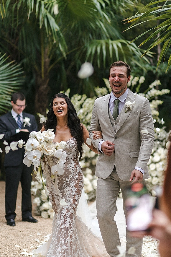 Wedding recessional with bride and groom walking down a flower-lined aisle, bride holding white orchid bouquet as petals fall in a palm garden