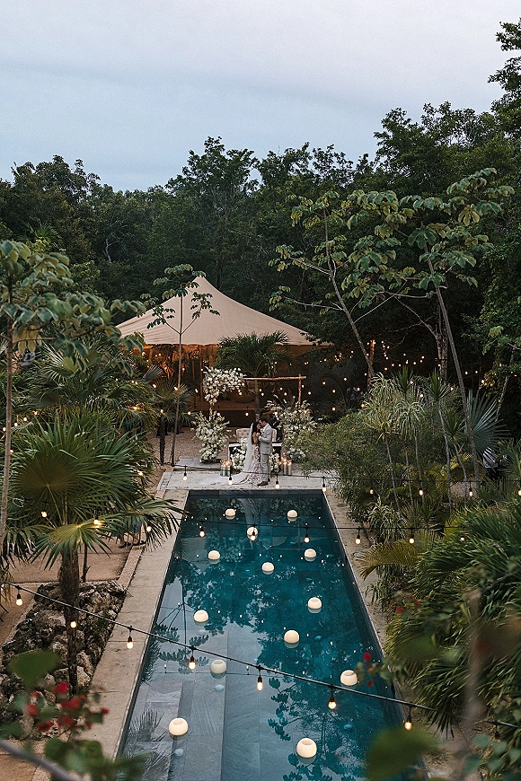Outdoor ceremony setup with a poolside wedding ceremony under string lights, wooden altar, aisle rug, and floating candles amid palms