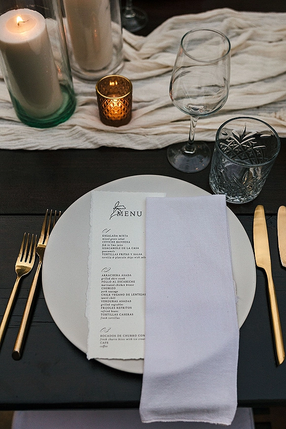 Wedding place setting with a wedding menu card on a white napkin, gold flatware, crystal glasses, and candlelight on a dark wood table