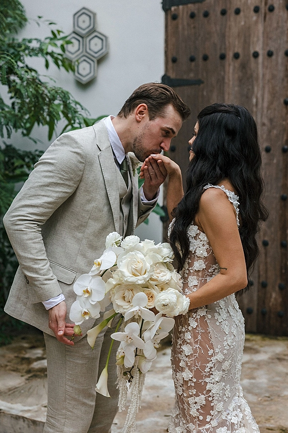 Couple portrait of groom kissing bride’s hand as she holds a cascading white orchid bouquet, posed by a rustic wooden door and stucco wall