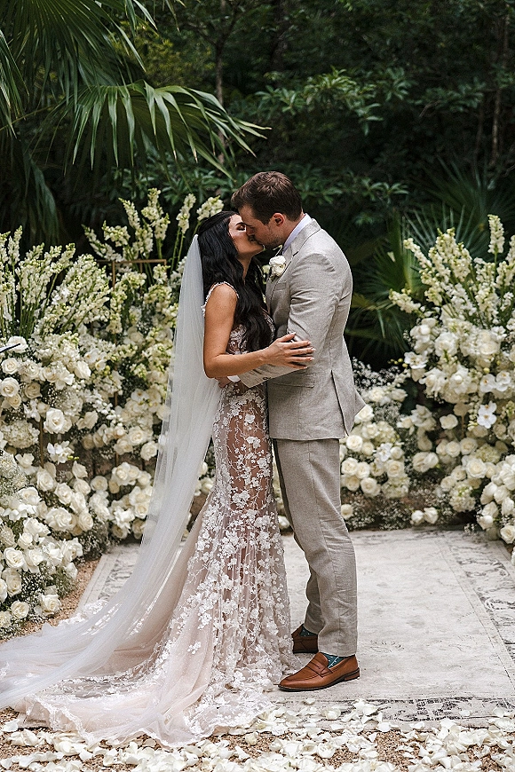 Wedding kiss portrait of bride and groom kissing, her lace dress and cathedral veil beside white flowers and rose petals amid tropical greenery