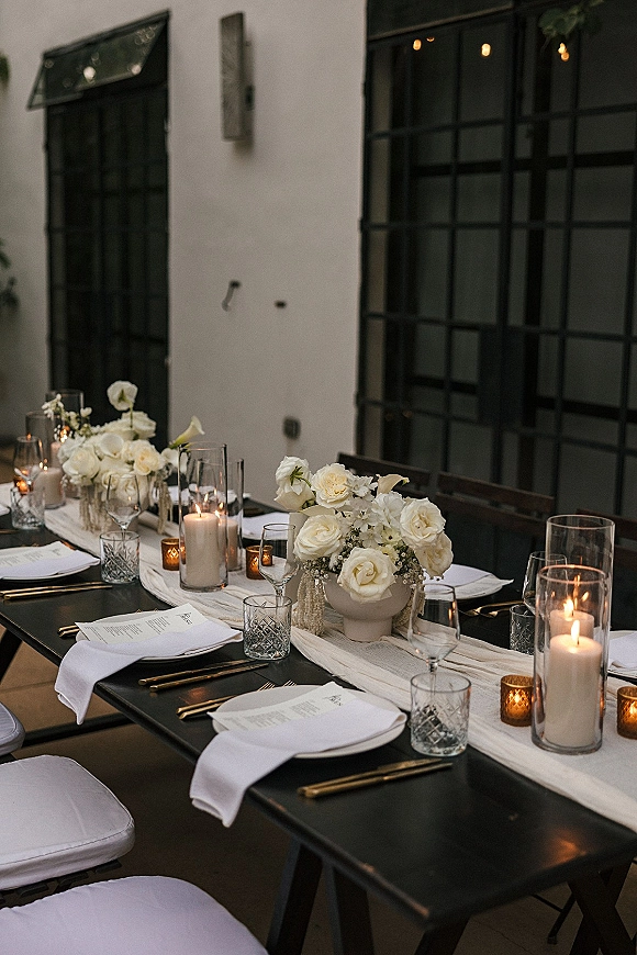 Reception tablescape with black and white wedding table styling, white rose centerpieces and glass hurricane candles on an outdoor patio under bistro lights