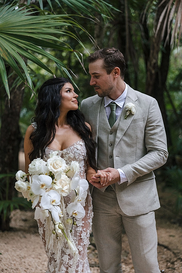 Couple portrait of bride and groom holding hands, her white orchid and rose bouquet beside a lace gown amid palm-filled garden path