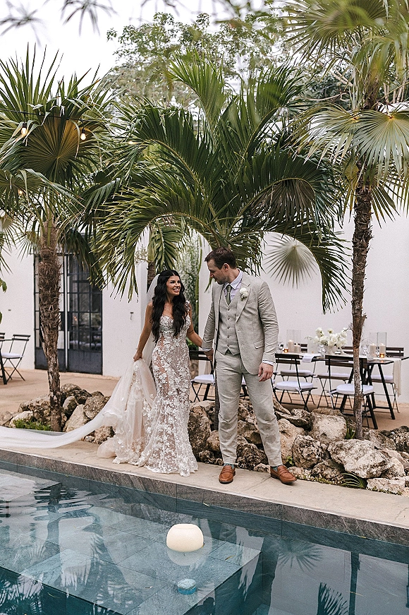 Couple portrait of bride and groom holding hands by a poolside patio, her lace dress and veil beside candlelit tables under string lights