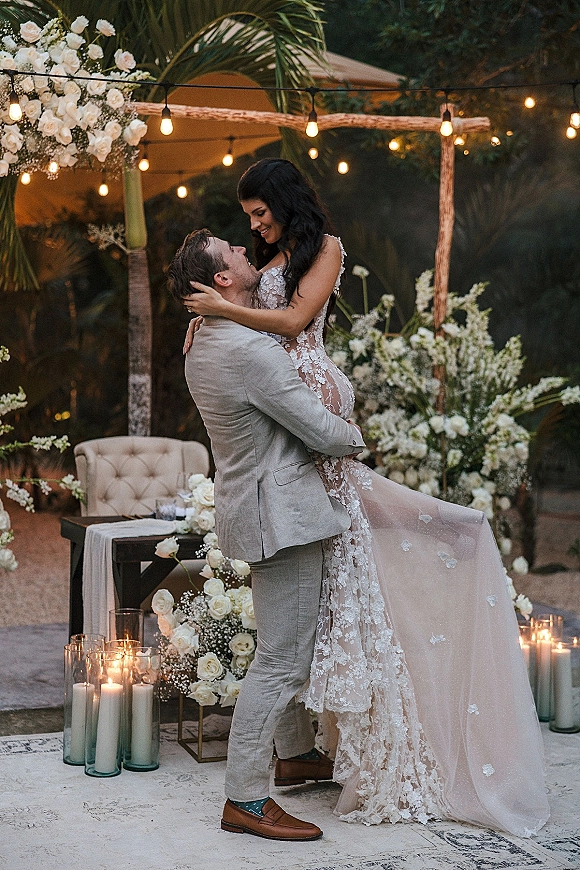 Couple portrait of groom lifting bride in a lace wedding dress with long train beneath a white floral arch with candles and string lights on a garden patio