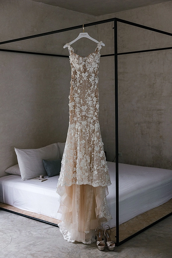 Wedding dress with lace appliqué hanging from a four poster bed frame, nude lining and spaghetti straps above bridal shoes on pillows