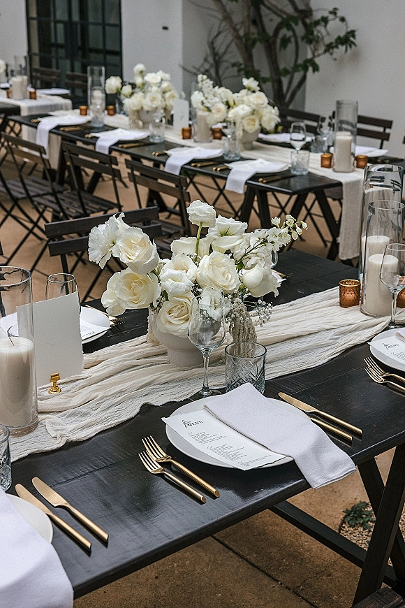 Reception tablescape with a long banquet table setting, white floral centerpieces and pillar candles on black tables beside a stucco wall with vines