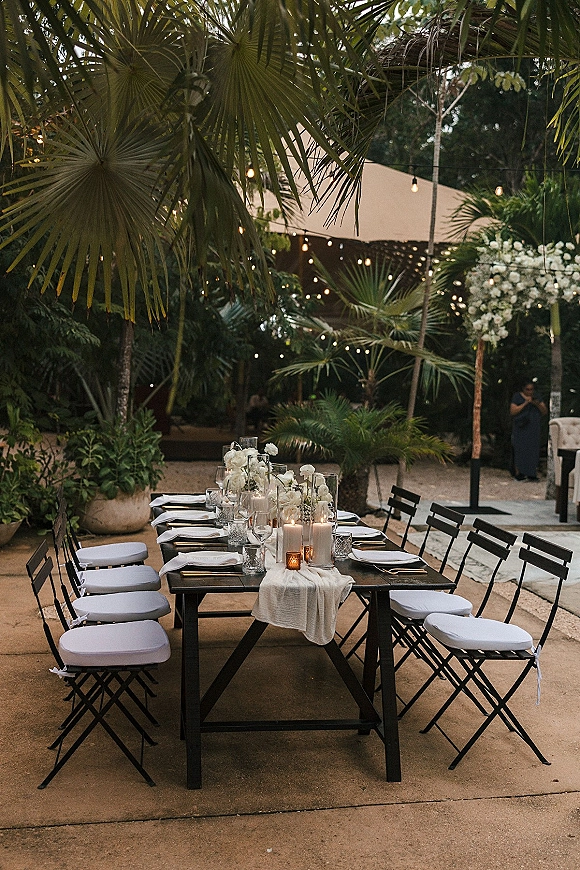 Reception tablescape with an outdoor reception table set on a long banquet table, candlelit hurricanes and white florals under string lights by palms