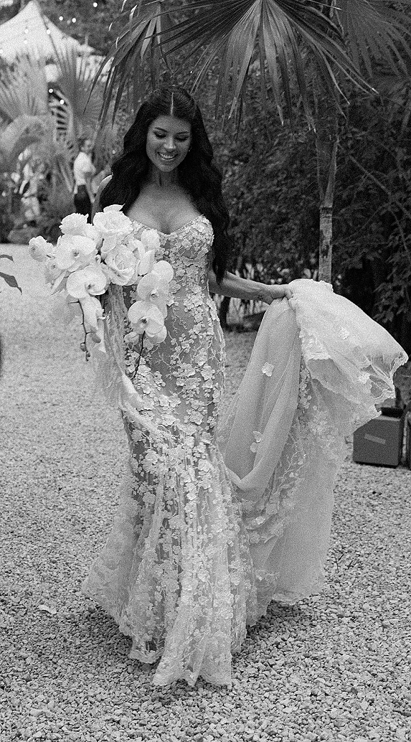 Bridal portrait of a bride on a garden path holding an orchid bouquet, wearing a lace mermaid gown with tulle overskirt and palm leaves behind