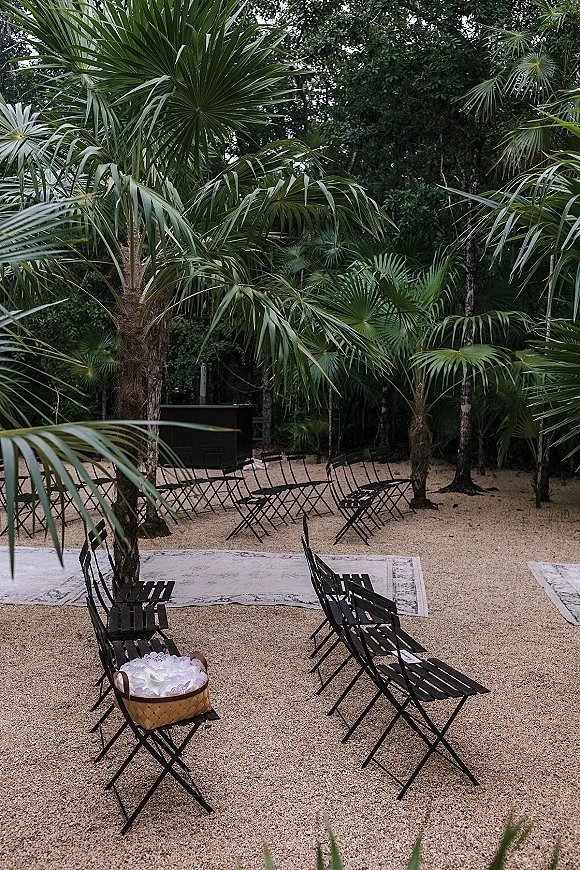 Ceremony setup for an outdoor wedding ceremony with black folding chairs, layered aisle rugs, and a petal basket on gravel amid palms