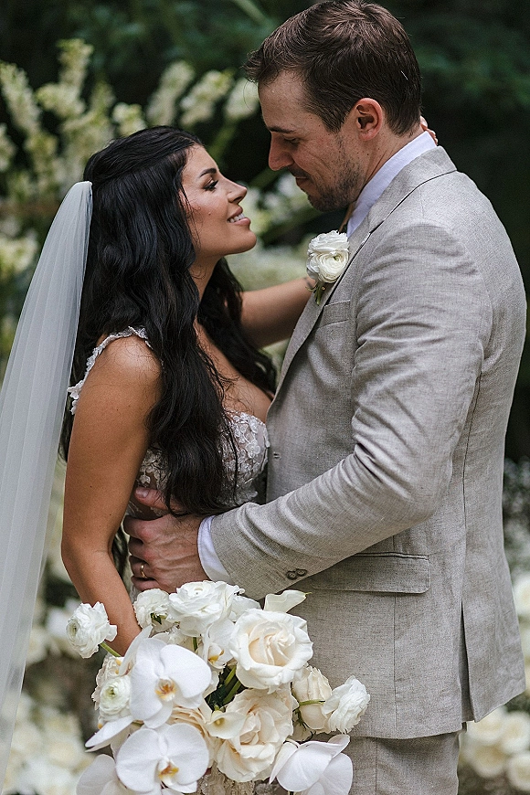 Couple portrait of bride and groom embrace, her lace dress and veil flowing as they gaze together before a white floral garden backdrop