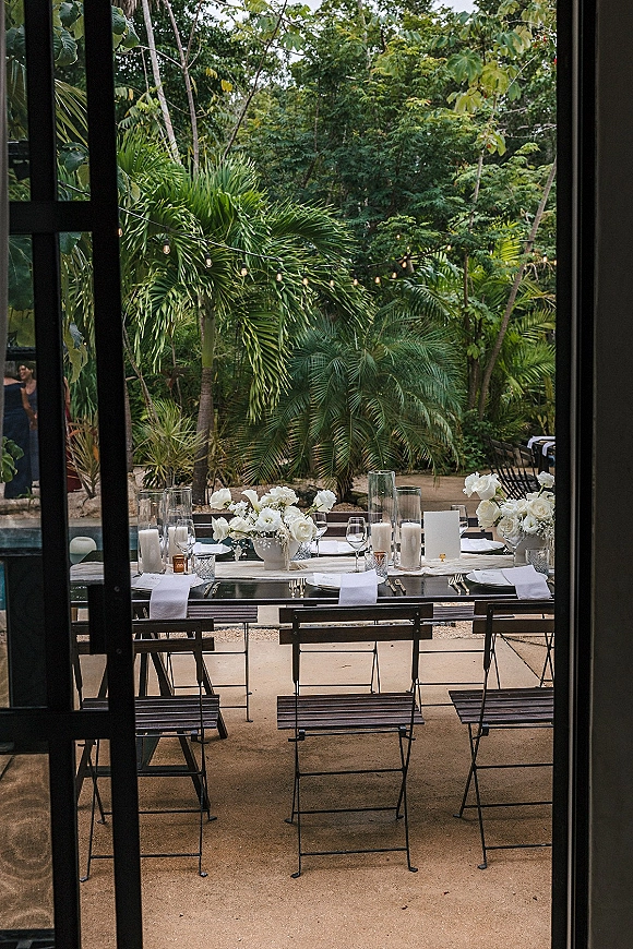 Reception tablescape with white floral centerpieces and candlelight, gold flatware and menus on a long poolside outdoor reception table under string lights