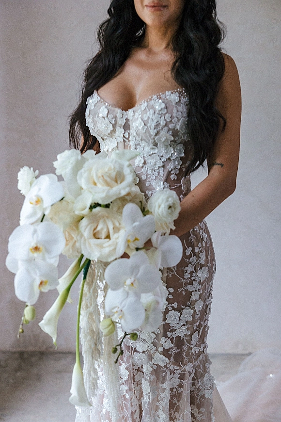 Bridal portrait of a bride in a lace wedding dress with floral appliques, holding a white rose and orchid bouquet against a neutral wall