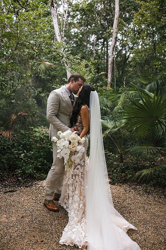 Wedding kiss portrait of bride and groom kissing, bride holding a cascading orchid bouquet with long veil on a gravel path in tropical forest