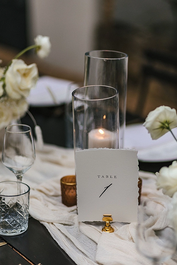 Reception tablescape with wedding table number card in a gold holder, white roses, glass cylinder candles, and glassware on a dark tabletop