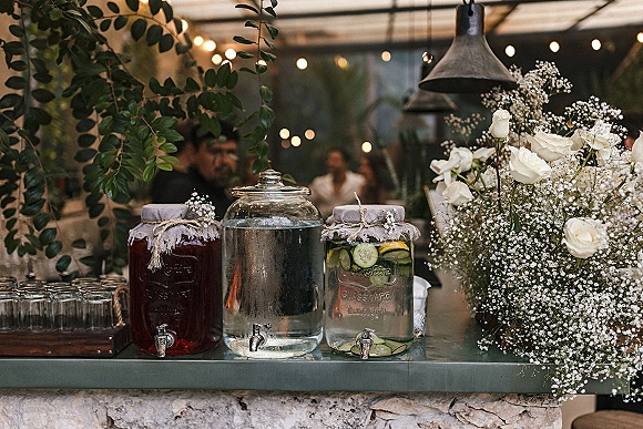 Wedding drink station with self serve beverage dispensers, cucumber lemon water, and stacked glasses under a tent canopy with string lights