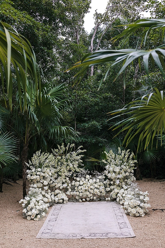 Ceremony backdrop with a floral ceremony backdrop of white roses and baby’s breath, ground arrangements and aisle rug amid tropical foliage