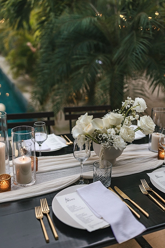 Reception tablescape with wedding table setting, white floral centerpiece and candlelight on a black runner beside a pool under string lights