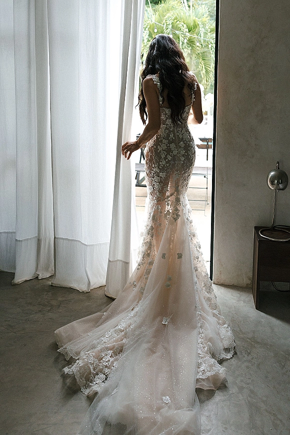 Wedding dress with illusion back and floral lace appliqué, seen from behind as the bride stands in an open doorway with palm trees outside
